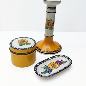 May include: A ceramic set consisting of a tall candlestick, a round lidded container, and an oval tray. Each piece features a mustard yellow base, white surfaces, and hand-painted floral motifs in yellow, pink, and blue with black geometric borders.