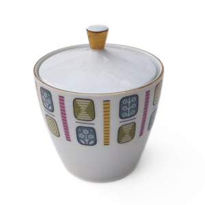 May include: A white ceramic sugar bowl with a lid and gold detailing. The bowl has a repeating pattern of colourful geometric shapes in blue, pink, yellow, and green. The lid has a gold knob.