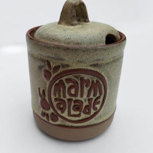May include: A ceramic marmalade jar with a lid and handle. The jar is light green with a brown rim and base. The word "marmalade" is embossed in brown on the side, along with a decorative design.