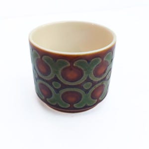 Può includere: Una tazza cilindrica in ceramica con esterno marrone, decorata con un motivo ripetuto di forme verdi e rosso-arancio. L'interno della tazza è beige chiaro. La tazza ha una finitura liscia e lucida.