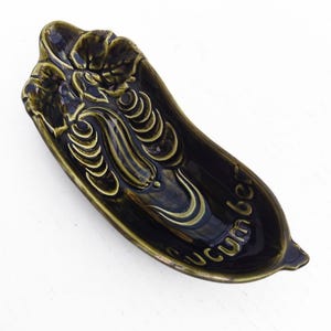 Può includere: Un piatto in ceramica verde scuro a forma di cetriolo con la parola "Cucumber" in corsivo. Il piatto presenta dettagli in rilievo di foglie e un motivo a cetriolo, ideale per servire o esporre.