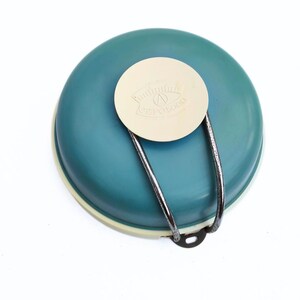 Puede incluir: Un cuenco turquesa con borde y tapa crema. La tapa tiene el texto "eepolooo". Un asa de metal plateado está unida. Ideal para uso en la cocina.