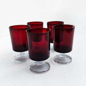 May include: A set of five ruby red wine glasses. Each glass has a cylindrical bowl and a clear, circular base. The glasses are arranged on a white surface, showcasing their rich color and design. Ideal for serving wine or other beverages.