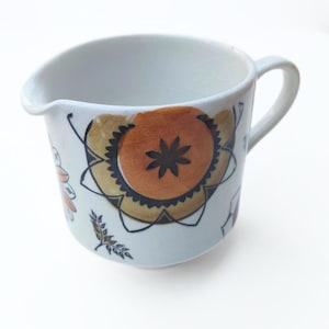 Può includere: Una lattiera in ceramica bianca con manico e beccuccio. La lattiera è decorata con un motivo floreale nei toni del marrone, arancione e nero. La lattiera è alta circa 10 cm.