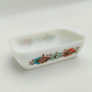 Op de afbeelding: Een rechthoekige, witte glazen schaal met een bloemmotief in teal, bruin en rood. De schaal heeft afgeronde hoeken en een iets verhoogde rand. Het bloemmotief herhaalt zich langs de zijkant van de schaal.