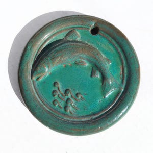 Puede incluir: Colgante redondo de cerámica turquesa con un diseño en relieve de un pez nadando. El pez está rodeado por un borde y tiene un pequeño agujero en la parte superior. El colgante tiene un aspecto rústico y artesanal.