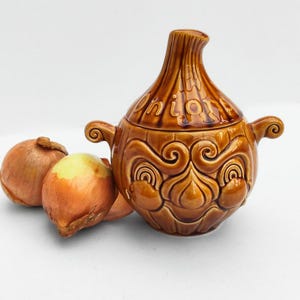 May include: A brown ceramic onion-shaped container with a lid, featuring the word "Onion" on top. The container has two handles and decorative swirls. Three onions are next to the container.