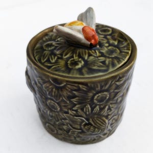May include: A ceramic jar with a lid, featuring a sunflower pattern and a decorative bee on top. The jar is a dark olive green color, with a textured surface. The bee has orange, white, and yellow accents.