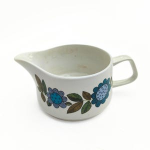 May include: White ceramic creamer with a handle and spout. The creamer is decorated with blue and teal flowers and brown leaves. The creamer is a vintage style and is suitable for serving cream or milk.