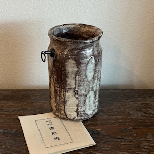 May include: A cylindrical ceramic vase with a textured brown and white finish, featuring a black handle and a slightly uneven rim. A small paper with Japanese text rests in front of the vase.
