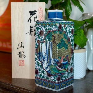 May include: A rectangular ceramic vase with a blue, green, and gold floral design, featuring a waterfall scene. The vase has a blue top and base, and is next to a wooden box with Japanese characters.