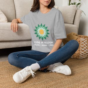 Könnte beinhalten: Ein graues T-Shirt mit einer Grafik einer stilisierten Blume mit grünen Blütenblättern und einem gelben Zentrum, das ein Codesymbol enthält. Unter der Blume steht der Text CODE IN BLOOM DEVELOPER / DESIGNER / CREATOR in weißen Buchstaben.