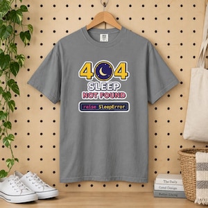 May include: A grey short-sleeved t-shirt featuring a graphic with the text 404 SLEEP NOT FOUND and raise SleepError. The design includes a dark blue circle with a yellow crescent moon and stars, surrounded by yellow and white text elements.