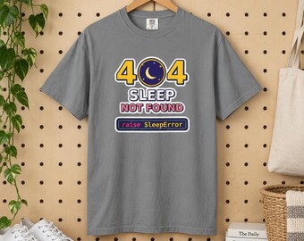 May include: A grey short-sleeved t-shirt featuring a graphic with the text 404 SLEEP NOT FOUND and raise SleepError. The design includes a dark blue circle with a yellow crescent moon and stars, surrounded by yellow and white text elements.