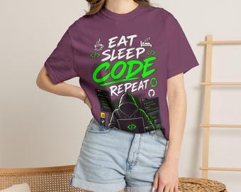 Op de afbeelding: Een pruimkleurig T-shirt met korte mouwen en de witte en felgroene tekst EAT SLEEP CODE REPEAT. De afbeelding bevat iconen van een koffiekopje, een slapend persoon en een figuur met een capuchon die op een laptop met codevensters werkt.