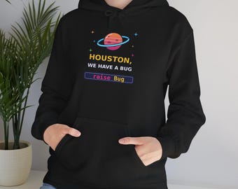 Pode incluir: Uma camisola preta com capuz apresentando um gráfico de um planeta com anéis, rodeado por pequenas estrelas coloridas. Abaixo do planeta, o texto diz HOUSTON, WE HAVE A BUG, seguido por uma caixa retangular com o código raise Bug.