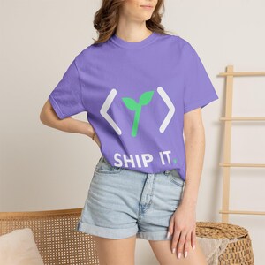 Puede incluir: Una camiseta de manga corta de color morado claro con un gráfico blanco de corchetes angulares que rodean un icono de brote verde. Debajo del gráfico, el texto dice SHIP IT. en letras mayúsculas blancas con un punto verde al final.