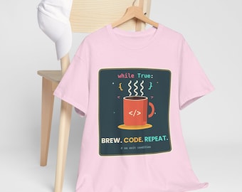 Pode incluir: Uma t-shirt de manga curta rosa claro com um gráfico de uma caneca de café vermelha com símbolos de código, vapor e o texto while True: BREW. CODE. REPEAT. # no exit condition. Um boné de basebol branco está pendurado no cabide de madeira.