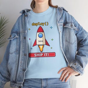 Puede incluir: Una camiseta azul claro con un gráfico de un cohete, el texto deploy() encima y una pancarta rectangular roja debajo con el texto SHIP IT!, rodeado de pequeños iconos de estrellas de colores.