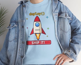 Op de afbeelding: Een lichtblauw t-shirt met een afbeelding van een raket, de tekst deploy() erboven en een rode rechthoekige banner eronder met de tekst SHIP IT!, omringd door kleine kleurrijke sterpictogrammen.