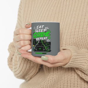 Peut inclure: Un mug en céramique gris avec le texte EAT SLEEP CODE REPEAT en lettres blanches et vertes. Sous le texte se trouve un graphique représentant une silhouette à capuche travaillant sur un ordinateur portable avec du code informatique.