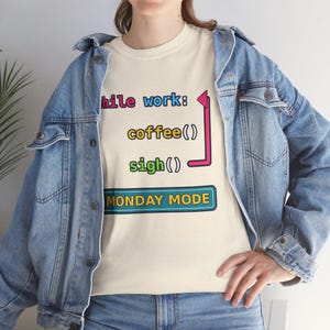 May include: A cream-colored t-shirt featuring a graphic with the text "while work: coffee() sigh()" in colorful letters, a pink bracket, and a blue rectangular box containing the text "MONDAY MODE" worn under a blue denim jacket.