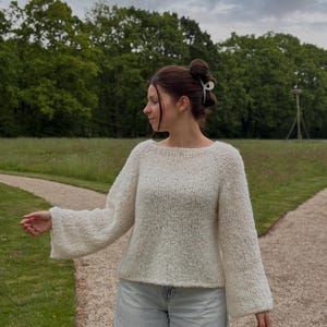 Puede incluir: Un suéter de punto texturizado color crema con mangas acampanadas anchas. El suéter se combina con unos vaqueros de lavado claro. El fondo presenta un parque con árboles verdes y un cielo nublado.