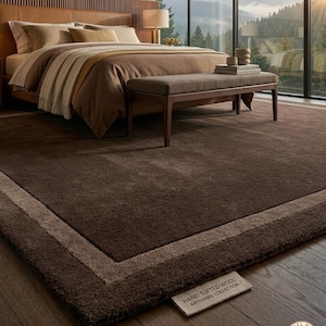 May include: A large rectangular area rug in a deep chocolate brown color with a lighter beige border. The rug features a soft, plush texture and includes a fabric tag that reads HAND TUFTED WOOL ARTISANAL COLLECTION and a circular logo with the letter R.