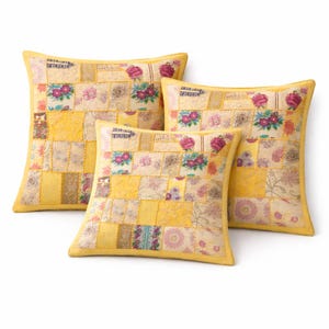Puede incluir: Tres cojines cuadrados decorativos con un diseño de patchwork. Los cojines tienen un fondo amarillo con estampados florales y geométricos en tonos rosa, morado y beige. Cada cojín tiene un borde amarillo.