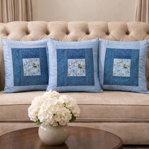 Peut inclure: Trois coussins carrés avec un fond bleu clair et un motif carré bleu foncé. Le centre de chaque coussin présente un carré bleu clair avec des détails floraux brodés. Les coussins sont disposés sur un canapé beige.