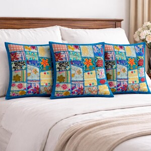 Puede incluir: Tres cojines cuadrados de patchwork con una vibrante mezcla de patrones florales y geométricos en tonos azules, amarillos y rojos. Cada cojín tiene un borde azul liso y está colocado sobre una cama blanca.