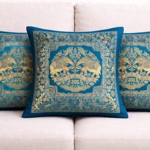 May include: Three teal square cushions with gold elephant and floral patterns, arranged on a white sofa. The cushions have a square shape and are made of a soft fabric. The design is symmetrical.