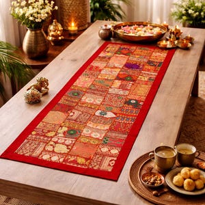 May include: A rectangular, patchwork-style table runner with a red border. The runner features a variety of patterned fabric squares in shades of red, orange, and gold. The runner is placed on a wooden table, with decorative items and food around it.