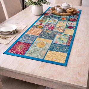 May include: A colourful patchwork table runner with a blue border, displayed on a light wooden table. The runner is decorated with various patterns and colours. A vase of flowers and a tea set are also on the table, adding to the decor.