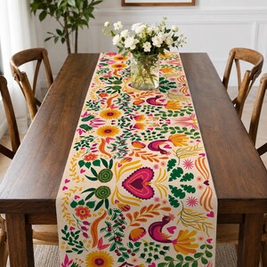 May include: A long rectangular table runner featuring a cream-coloured background with a vibrant, repeating folk-art pattern of stylised flowers, green leaves, orange vines, and purple birds, centred with a clear glass vase of white flowers.