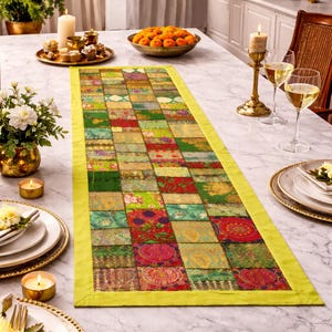 Op de afbeelding: Een kleurrijke patchwork tafelloper met een heldergele rand. De loper heeft een verscheidenheid aan vierkanten van stof met patronen in groen, rood en goud. De loper wordt getoond op een marmeren tafel met kaarsen en bloemen.