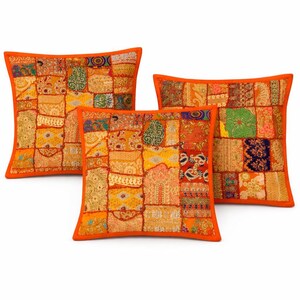 Puede incluir: Tres cojines cuadrados con diseños de patchwork en tonos naranja, amarillo y rojo. Cada cojín presenta una vibrante mezcla de patrones y texturas, con un borde naranja liso. Los cojines están dispuestos sobre un fondo blanco.