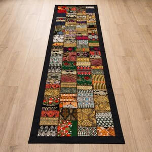 May include: A rectangular patchwork rug with a black border. The rug is composed of numerous fabric squares in various colors and patterns, including floral, geometric, and paisley designs. The rug is placed on a light-colored wooden floor.