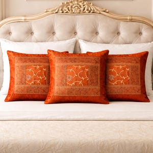 May include: Three square orange throw pillows with a floral design and decorative border are arranged on a bed. The pillows have a soft, velvety texture and are placed against white pillows and a decorative headboard.