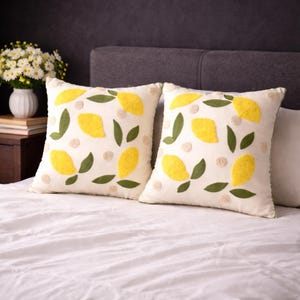 Può includere: Due cuscini quadrati con sfondo color crema. Ogni cuscino presenta limoni gialli ricamati, foglie verdi e piccoli pois beige. I cuscini sono appoggiati su un copriletto bianco.