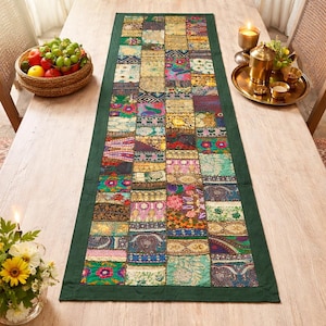 Könnte beinhalten: Ein rechteckiger Patchwork-Tischläufer mit dunkelgrünem Rand. Der Läufer zeigt eine Vielzahl farbenfroher Stoffquadrate mit floralen und geometrischen Mustern. Er liegt auf einem hellen Holztisch, mit einem Obstkorb und einem Messingtablett mit Teetassen.