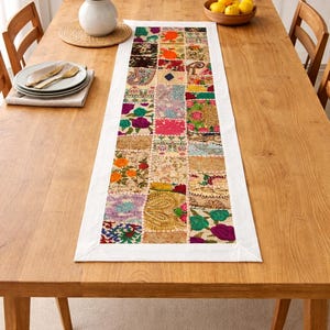 May include: A colourful patchwork table runner with a white border, displayed on a wooden table. The runner is made up of various fabric squares. A vase of flowers and a bowl of lemons are also visible.
