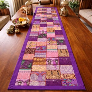 May include: A patchwork table runner with a vibrant mix of colors, including purple, pink, and gold. The runner features a geometric pattern of squares and rectangles, with a solid purple border. The runner is placed on a wooden table.