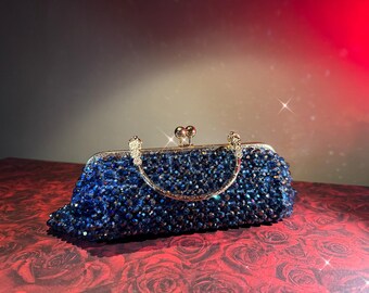 May include: A blue beaded clutch with a gold-colored handle and clasp. The rectangular bag is covered in small, dark blue beads, and the handle arches over the top. The clasp is a small, round, gold-colored metal piece.