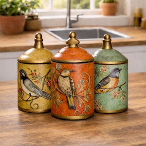 May include: Three decorative ceramic jars with lids. Each jar is painted with a different bird and floral design. The jars are in shades of yellow, orange, and green, with gold accents. The jars are sitting on a wooden surface.