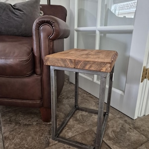 May include: A small side table with a rustic design. The tabletop is made of thick, light brown wood, showcasing the natural grain. It rests on a sturdy, square metal frame. The table is placed next to a brown leather armchair.