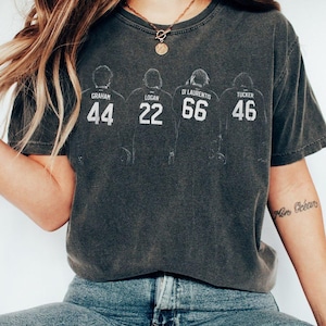 Op de afbeelding: Donkergrijs t-shirt met vier figuren en namen: Graham 44, Logan 22, Di Laurentis 66 en Tucker 46. Het witte ontwerp heeft een versleten, vintage uitstraling. Het t-shirt is een casual unisex stijl.