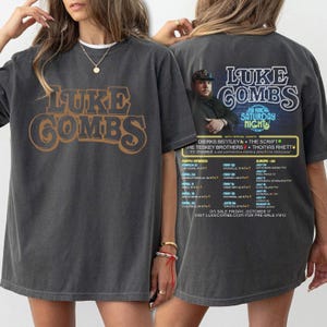 May include: Dark gray t-shirt featuring the text "LUKE COMBS" in brown and gold lettering on the front. The back of the shirt has a concert graphic with the text "KINDA SATURDAY NIGHT" and a list of tour dates.