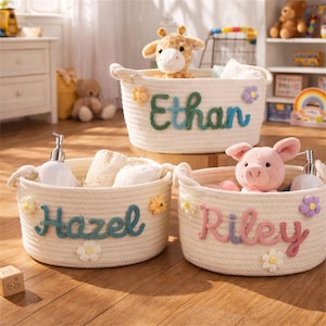 May include: Three off-white rope baskets with handles, each with a child's name in colorful letters. The baskets are decorated with fabric flowers and contain toys and toiletries. The names are Ethan, Hazel, and Riley.
