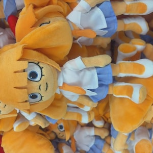 May include: A pile of plush dolls with yellow hair, blue eyes, and orange beaks. The dolls wear white shirts and blue skirts. Some dolls have red bows. The dolls are made of soft, plush material.